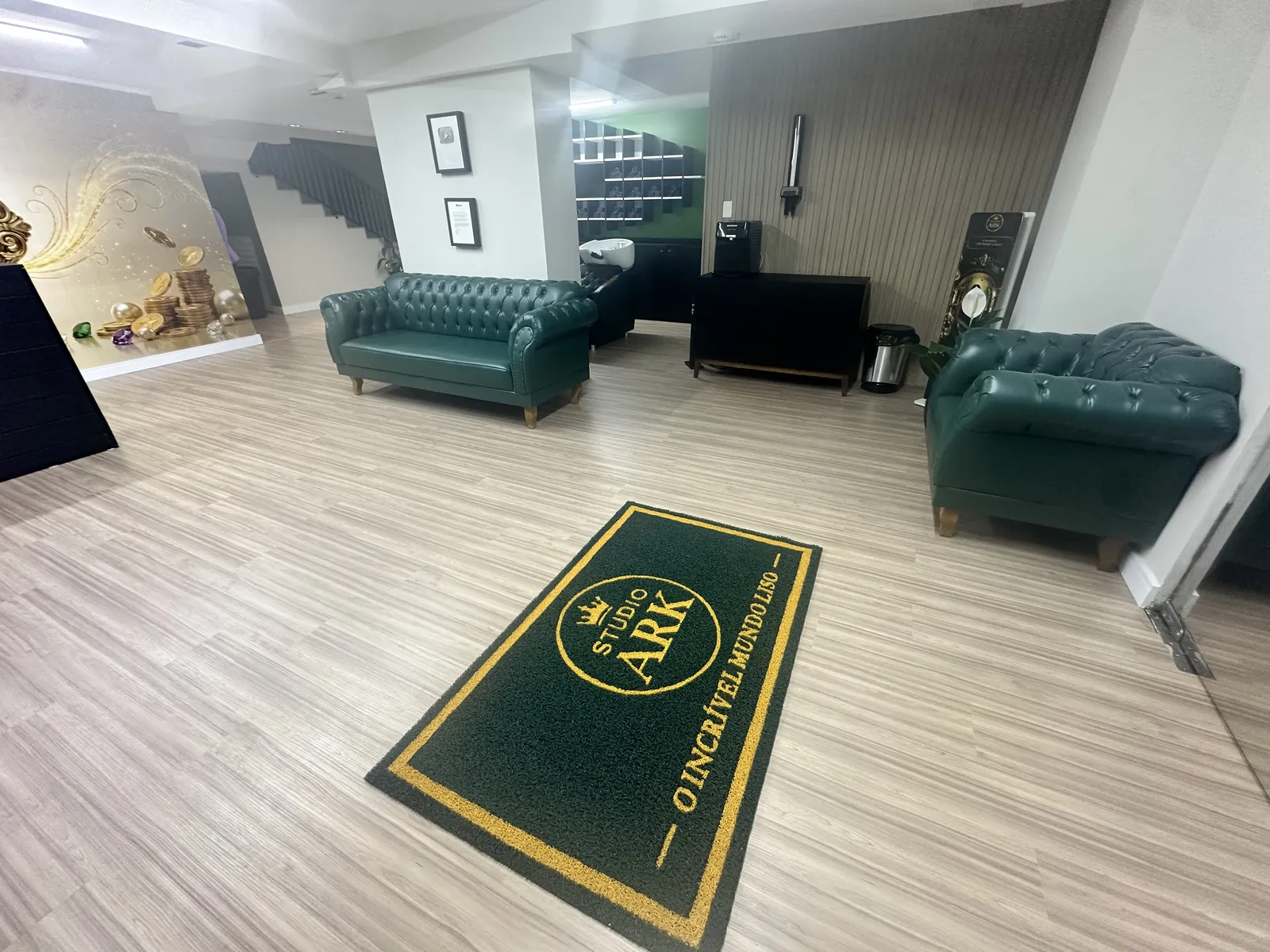 Outro ângulo da recepção do Studio Ark, com sofá e poltrona verdes, painel de madeira e tapete da marca.